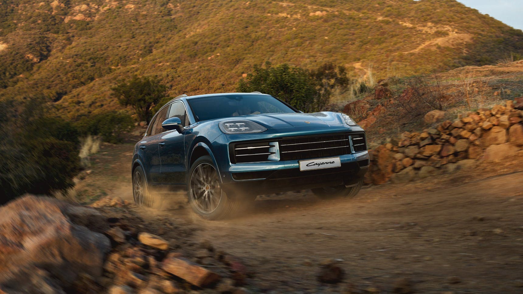 Montego Blue Metallic Cayenne SUV driving on a dusty, rocky path with hills in the background under a clear sky.